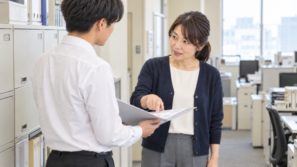 若手男性社員が資料を示し、先輩女性社員が相手の目を見て説明する様子