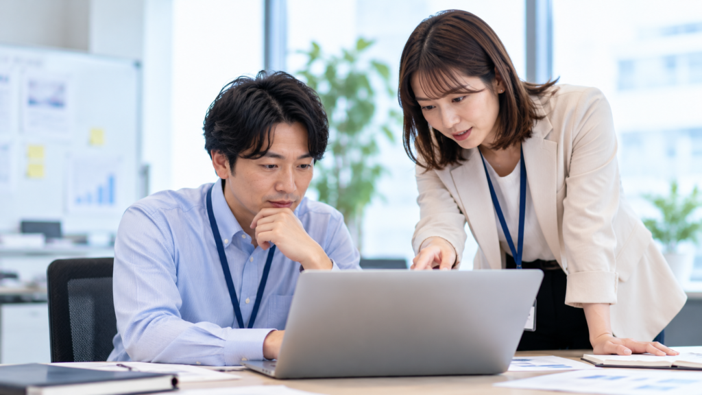 女性社員が男性社員のノートPC画面を指し、生成AIの利用について説明する様子